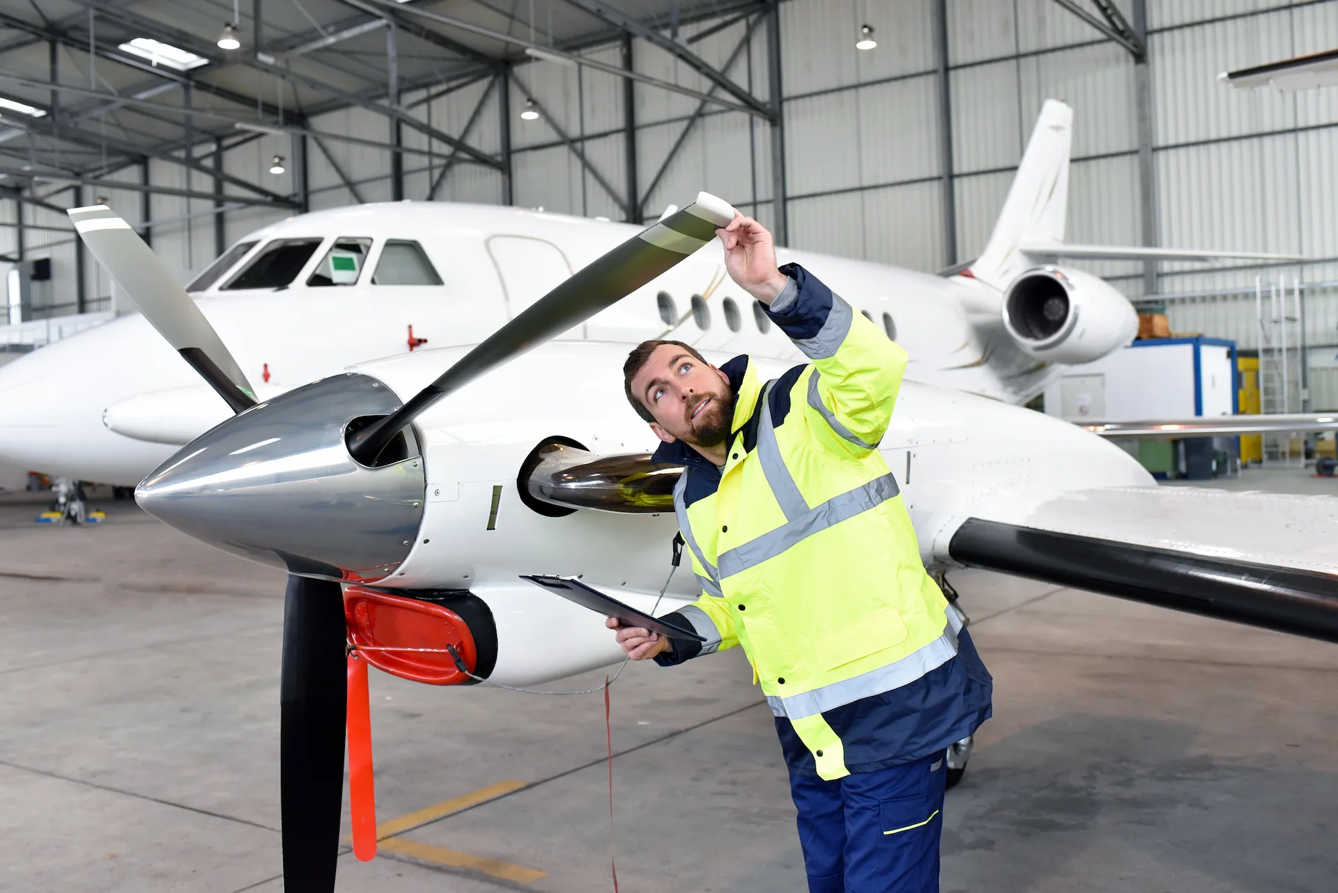 A mechanic in a high-visibility jacket inspects the propeller of an airplane inside a hangar, holding a clipboard and looking closely at the blade. Another jet aircraft is visible in the background.