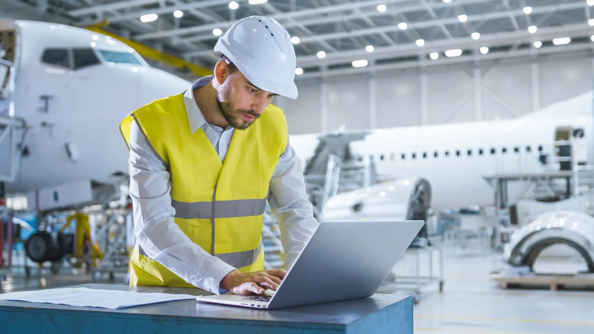 A man in a safety vest and hard hat analyzes data on a laptop in an aircraft maintenance facility. An airplane is visible in the background.